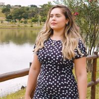 A imagem mostra Andressa em um parque. Ela é uma mulher, branca, de cabelos loiros longos e usa um vestido preto de bolinhas brancas.